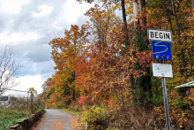 A portion of the Saranac River Trail, with a stone retaining wall to the left and bright fall foliage on the right. In the foreground there is a trail sign that says "BEGIN."